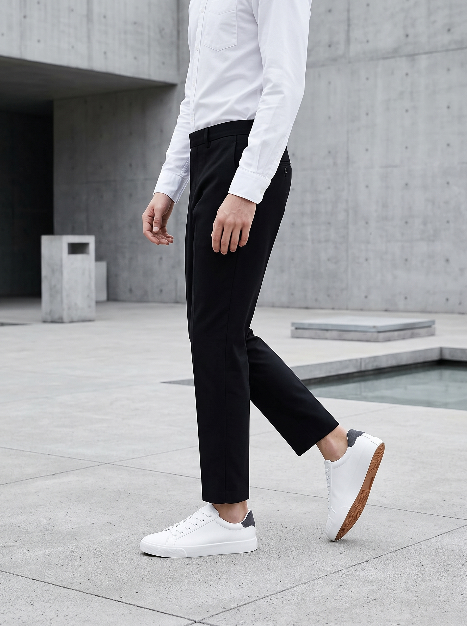A slim-fit menswear model wearing minimalist white PU leather tennis sneakers and black trousers in a modern urban setting for a spring lookbook.